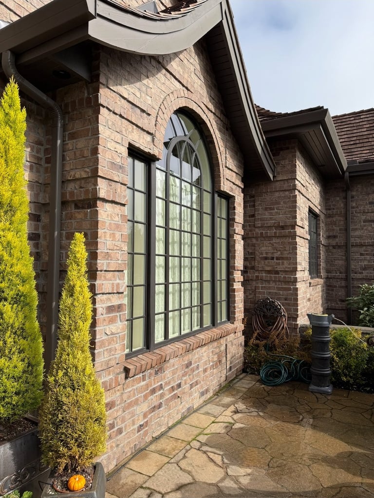 Brick residential building exterior with large green-framed arched window, manicured shrubs, and stone patio entrance.