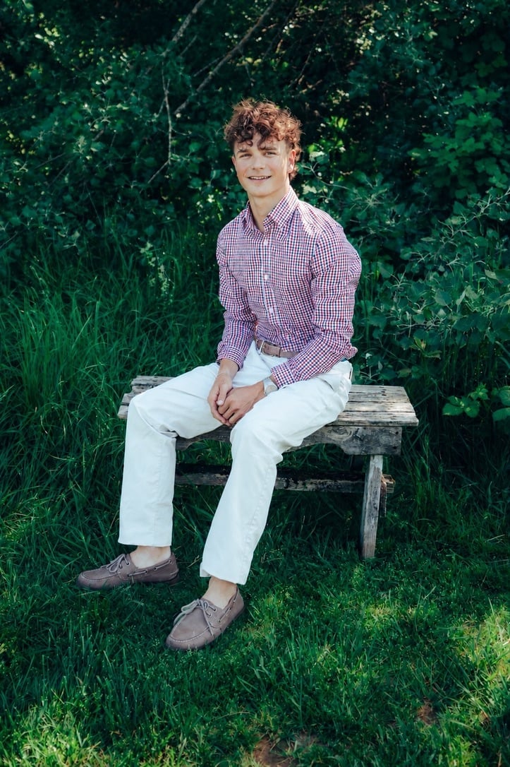 Young man with curly auburn hair sitting on a wooden bench, wearing a purple checkered shirt and white pants, surrounded by green foliage