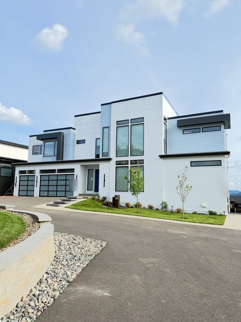 Modern white and gray residential house with geometric architecture, glass panels, driveway, landscaping, and mountain view in background